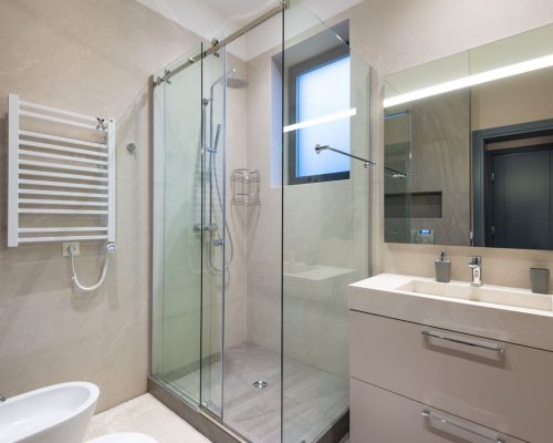 Shower cabin with glass walls near mirror hanging on wall above sink in modern minimalist styled bathroom